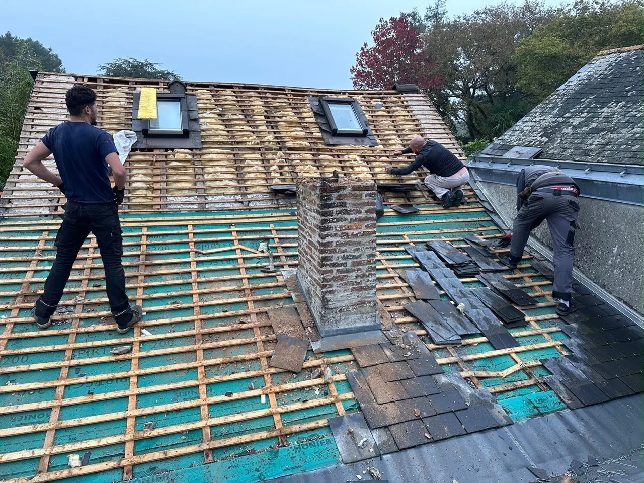 Artisans intervenant sur une toiture en ardoise autour de deux fenêtres de toit et d’une cheminée.